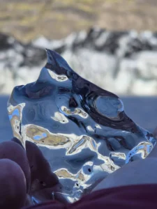 Close-up of a clear ice chunk held in a hand, reflecting light with a glacier blurred behind.