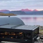 Modern Hvammsvík Hot Springs building on the shoreline, overlooking a calm fjord and sunset-glowing mountains.