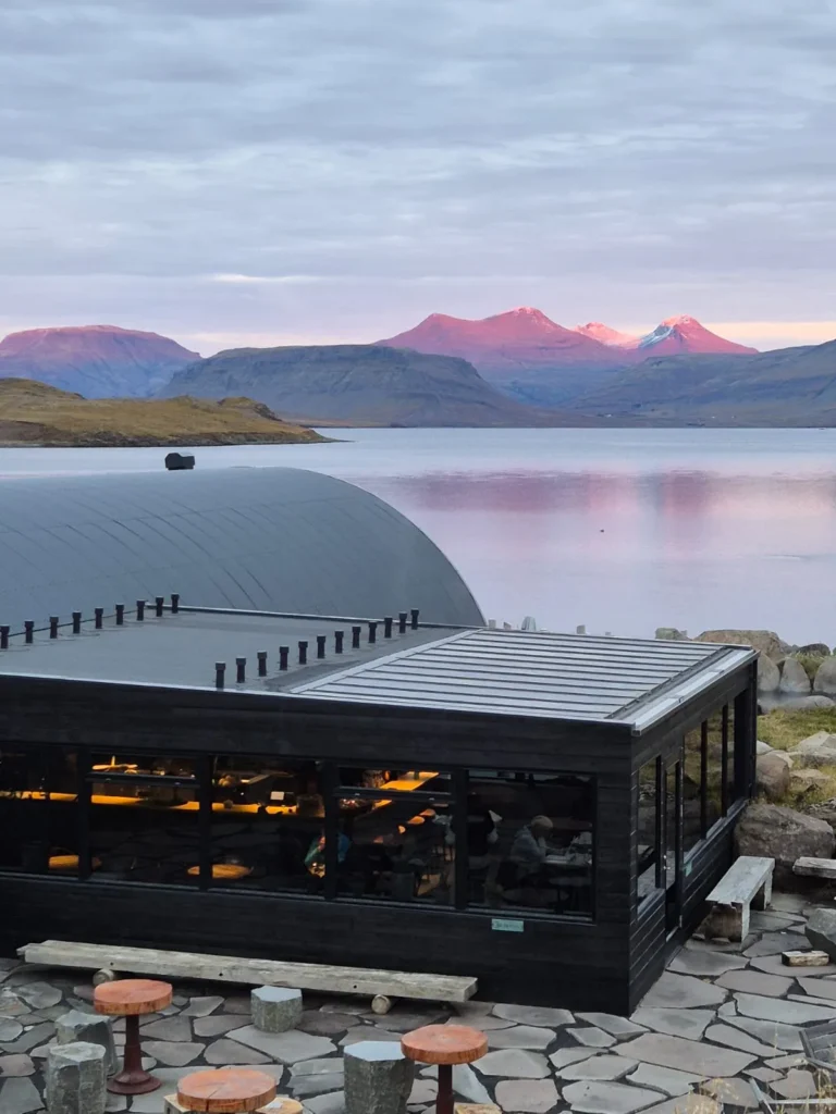 Modern Hvammsvík Hot Springs building on the shoreline, overlooking a calm fjord and sunset-glowing mountains.