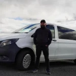 Otto, wearing a black cap and puffer vest, poses beside a white van under cloudy Icelandic skies.