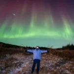 Person standing with arms outstretched under green northern lights in Iceland.