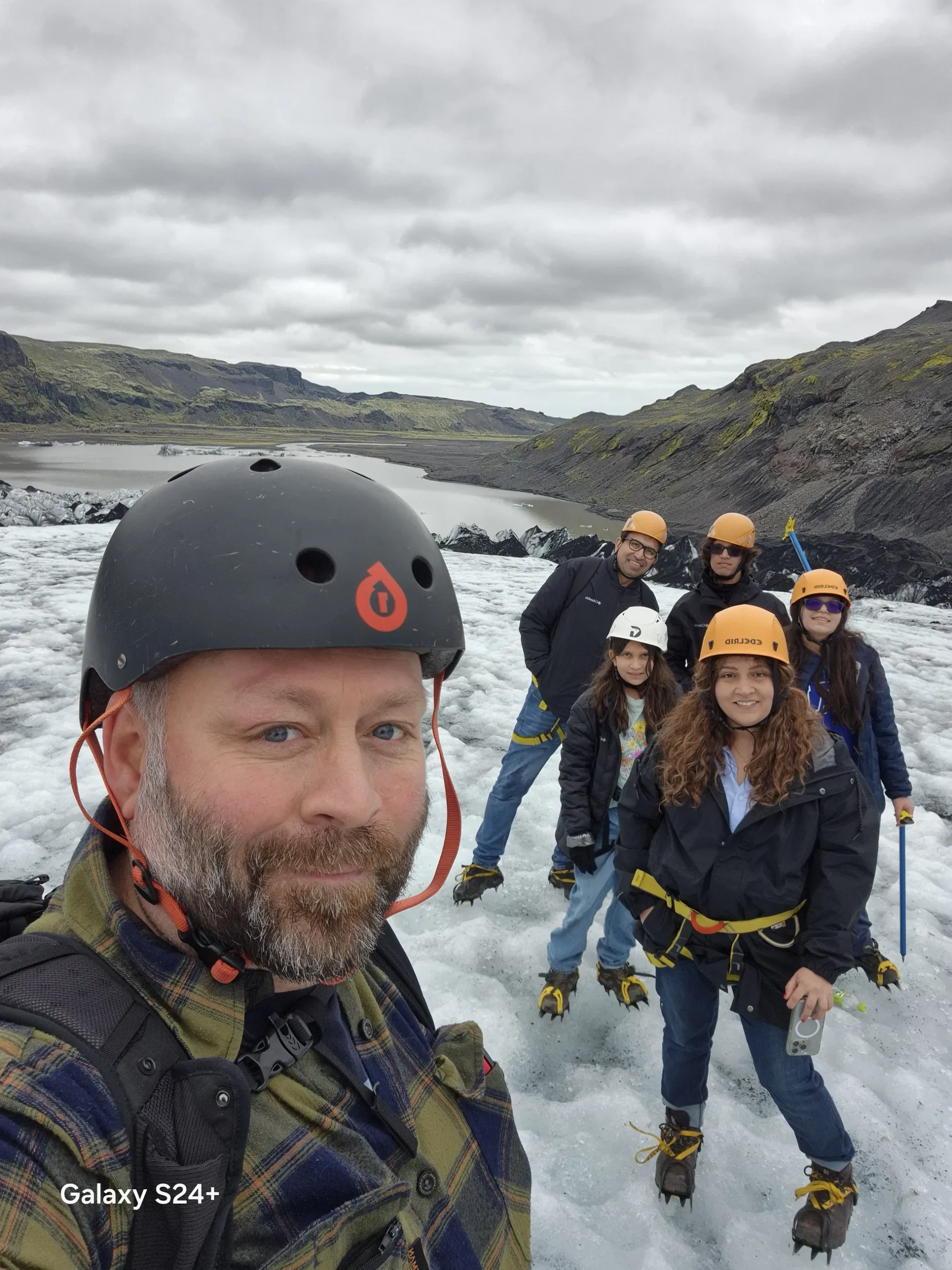 Smiling hikers in helmets and safety harnesses pose on a glacier with a glacial lagoon and rugged mountains behind them.