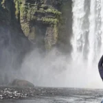Traveler forms a heart with their hands in front of a misty Iceland waterfall.