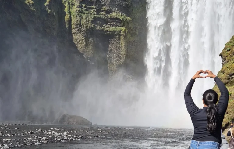Traveler forms a heart with their hands in front of a misty Iceland waterfall.