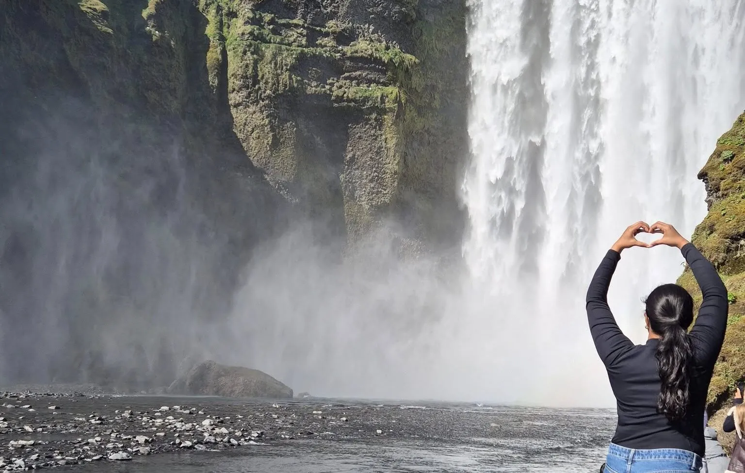Traveler forms a heart with their hands in front of a misty Iceland waterfall.