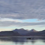 Calm Icelandic fjord reflecting distant mountains beneath a wide, cloudy sky.