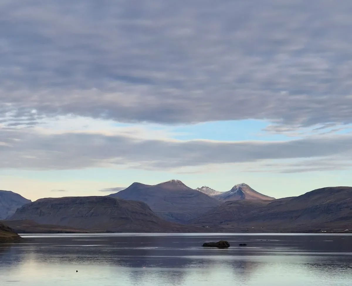 Calm Icelandic fjord reflecting distant mountains beneath a wide, cloudy sky.