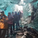 Tour group wearing helmets and winter gear walks through a blue ice cave in Iceland, with sunlight shining through the glacier walls.