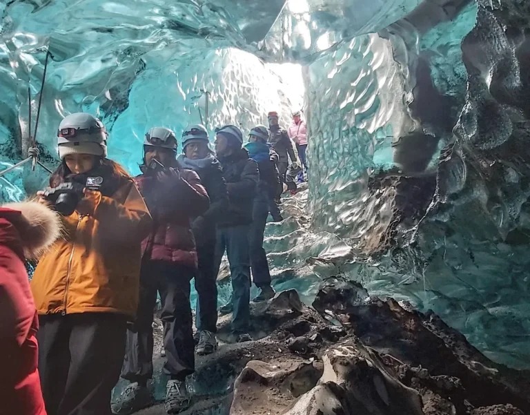 Tour group wearing helmets and winter gear walks through a blue ice cave in Iceland, with sunlight shining through the glacier walls.