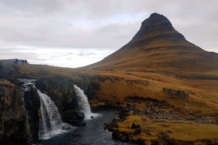 Waterfall and stream beneath Kirkjufell mountain in Iceland under a cloudy sky.