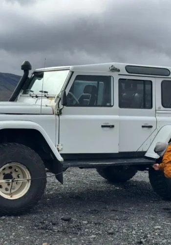 A person crouches beside a white off-road 4x4, adjusting a rear tire on a gravel track with mountains and cloudy skies behind.