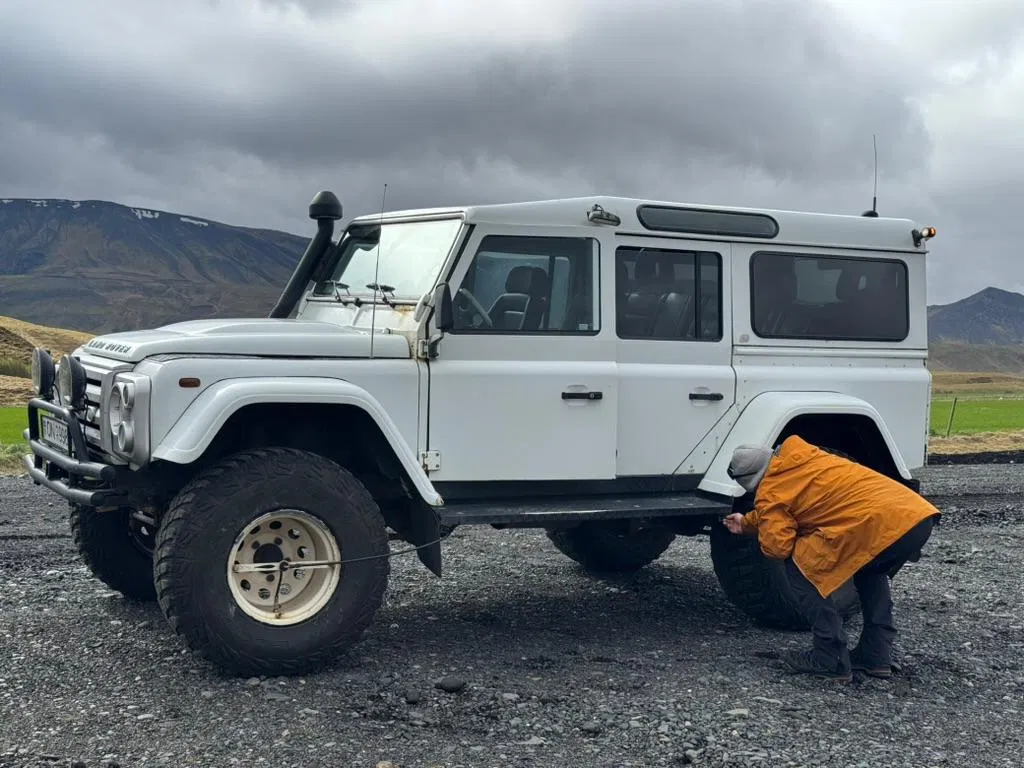 A person crouches beside a white off-road 4x4, adjusting a rear tire on a gravel track with mountains and cloudy skies behind.