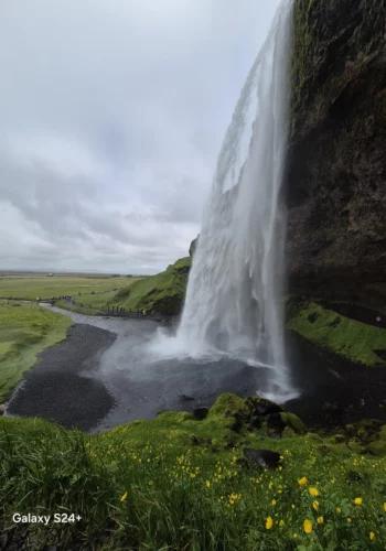 A tall Icelandic waterfall plunges into a dark rocky pool, with green hills and a walking path in the distance.