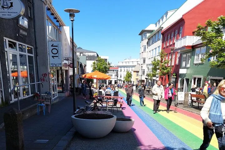 Busy Reykjavik street lined with colorful buildings, outdoor café seating, and a rainbow-painted road under a clear blue sky.