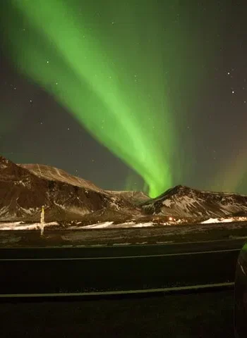 Green northern lights over snowy mountains beside an Icelandic road at night.
