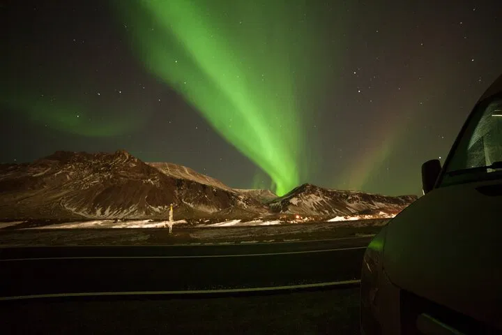 Green northern lights over snowy mountains beside an Icelandic road at night.