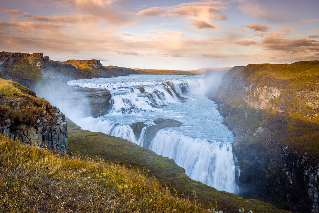 Gullfoss waterfall pours over broad stepped cliffs into a deep canyon, with green mossy slopes and warm evening light illuminating the landscape.