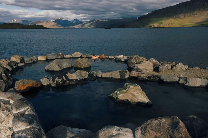 Hvammsvík Hot Springs pool bordered by rocks beside the fjord, with mountains and dramatic clouds in the distance.
