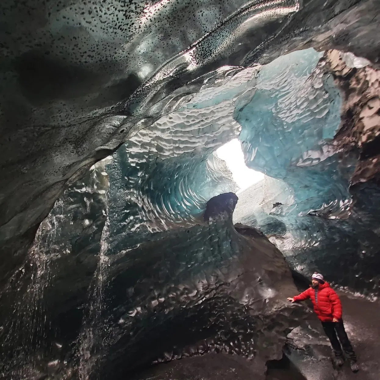 Inside an Icelandic ice cave, rippled blue-and-black ice arches overhead while a person in a red jacket stands on dark volcanic sand near the cave wall.