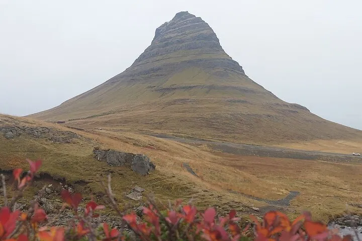 Kirkjufell mountain on Iceland’s Snæfellsnes Peninsula rises above grassy slopes, with red autumn shrubs in the foreground.