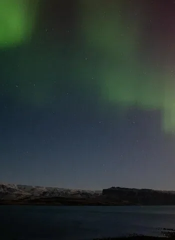 Northern lights glowing over a calm bay and dark hills in Iceland.