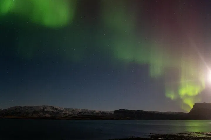 Northern lights glowing over a calm bay and dark hills in Iceland.