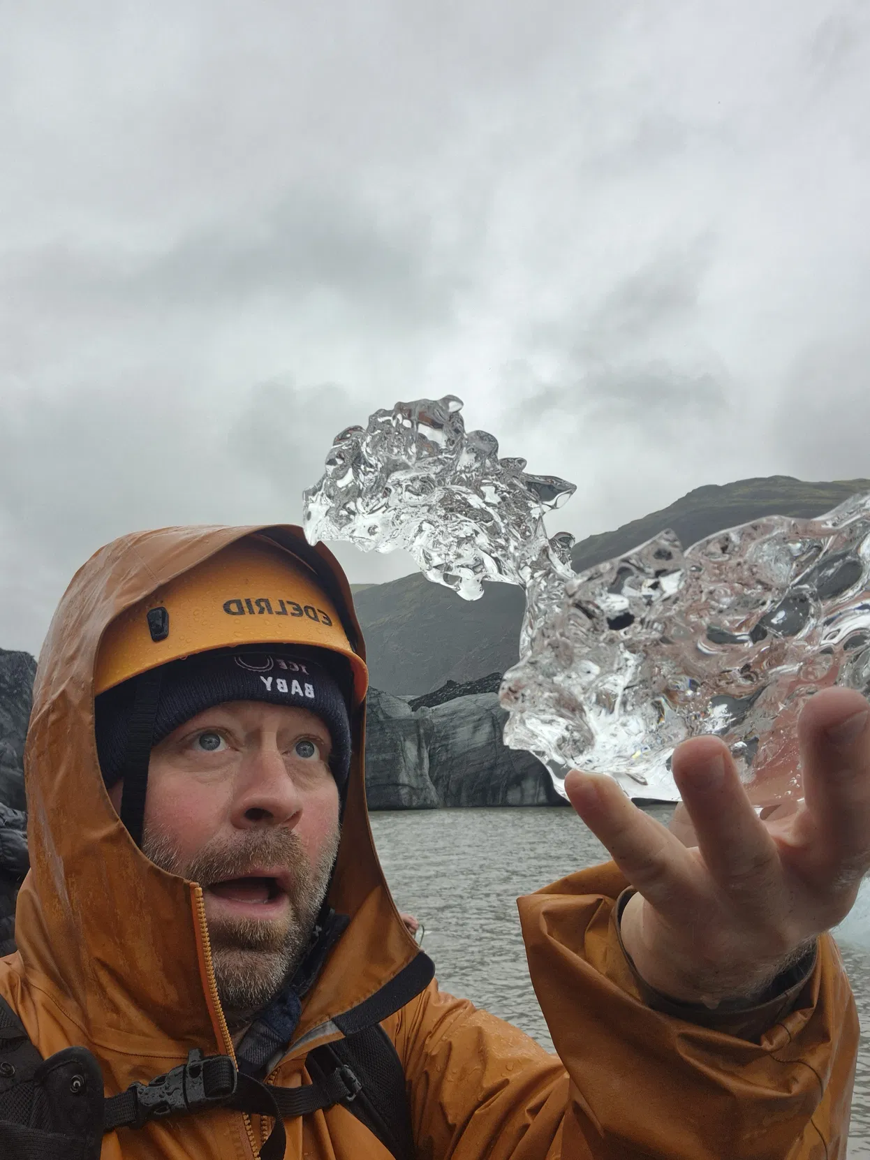 Otto in an orange rain jacket looks amazed while holding a crystal-clear piece of glacial ice in South Coast Iceland.