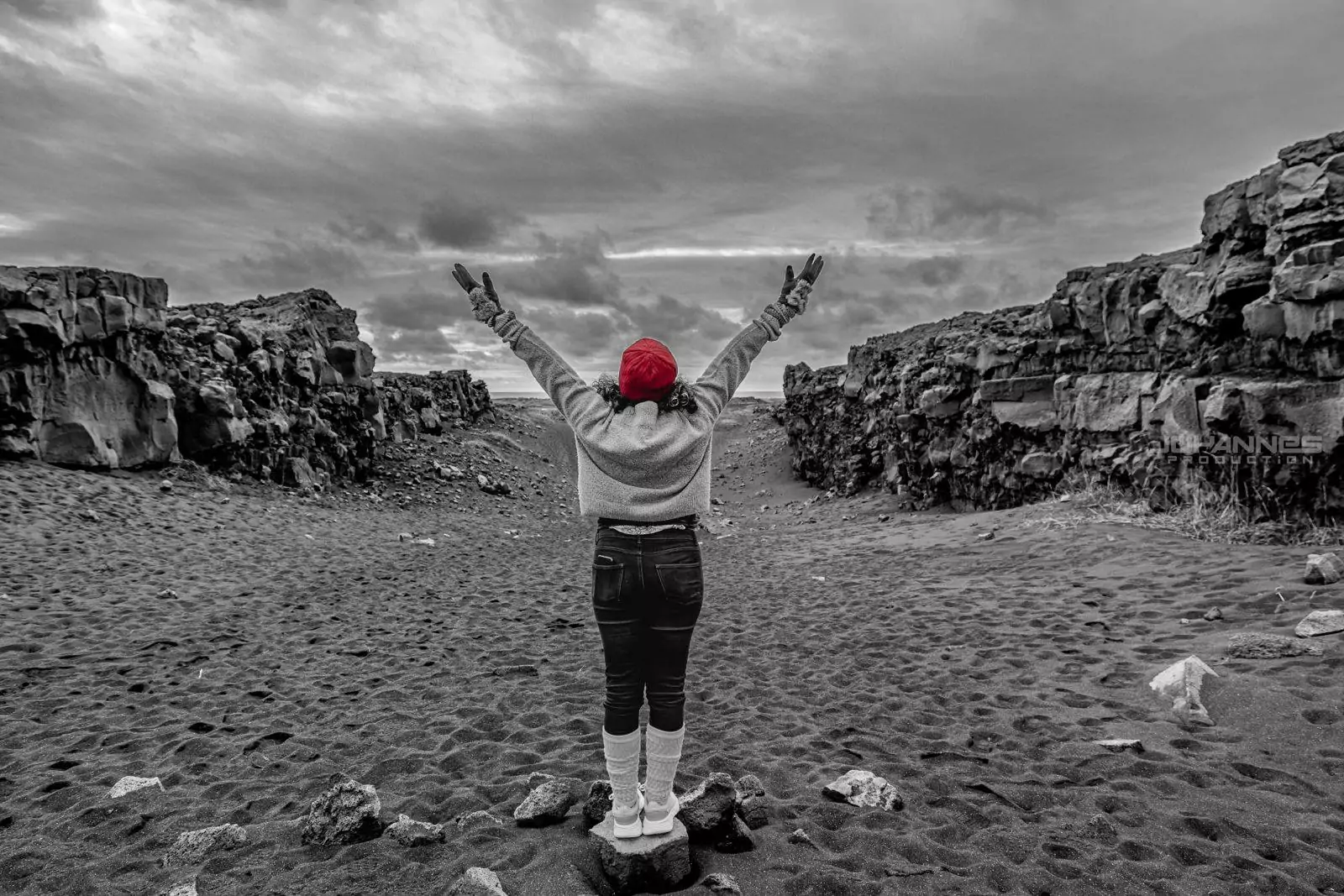 Person in a red beanie stands on black sand between rocky cliffs in Iceland, arms raised toward a cloudy sky.