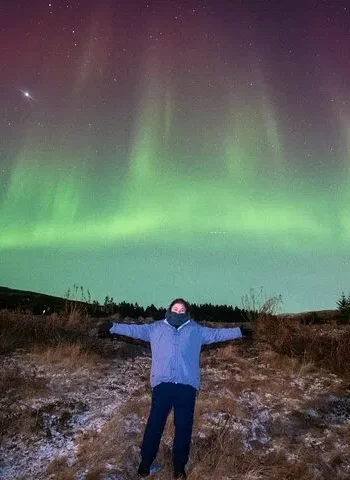 Person standing with arms outstretched under green northern lights in Iceland.