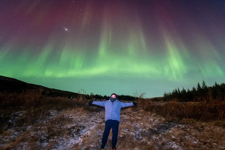 Person standing with arms outstretched under green northern lights in Iceland.