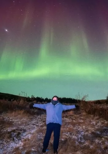 Person standing with arms outstretched under green northern lights in Iceland.