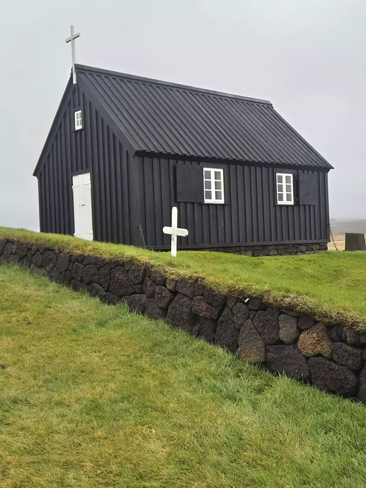 Small black wooden church with a white cross on the roof sits on a grassy hill in Iceland under an overcast sky.