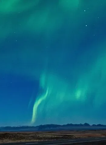 Soft green aurora curtains ripple across a starry sky over a wide Icelandic landscape with a mountain range on the horizon.