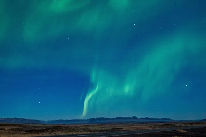 Soft green aurora curtains ripple across a starry sky over a wide Icelandic landscape with a mountain range on the horizon.