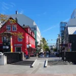 Sunny Reykjavik street scene with a bright red building, pedestrians in a plaza, and modern buildings under a clear blue sky.