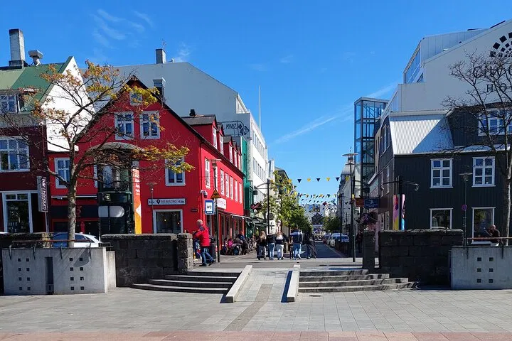 Sunny Reykjavik street scene with a bright red building, pedestrians in a plaza, and modern buildings under a clear blue sky.