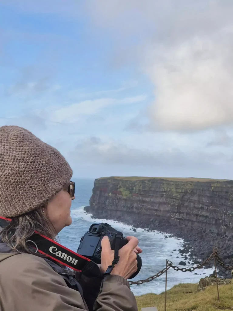 Traveler in a knit hat and sunglasses holds a camera while looking out over Iceland’s rugged sea cliffs and crashing waves.