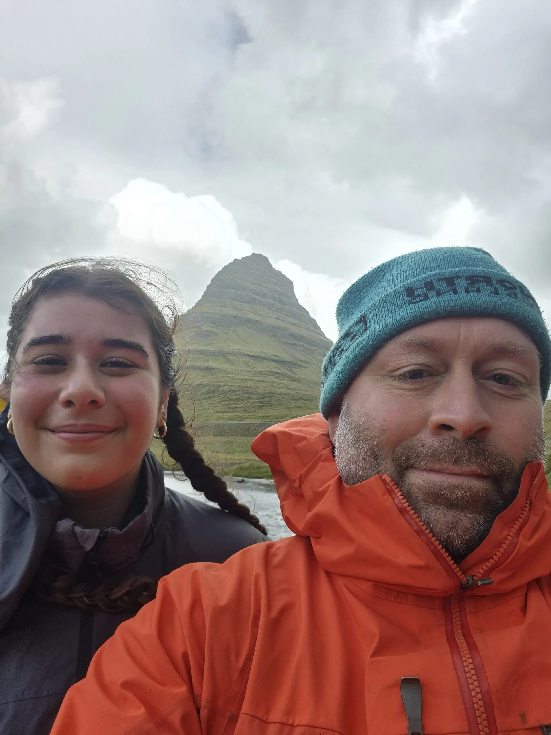 Two travelers smile for a selfie on Icelands Snæfellsnes Peninsula with the cone-shaped Kirkjufell mountain rising behind them under cloudy skies.
