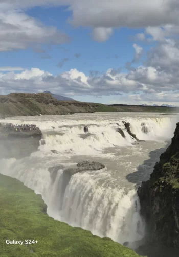 Wide Icelandic waterfall thundering over a rocky canyon under a blue, cloud-filled sky.