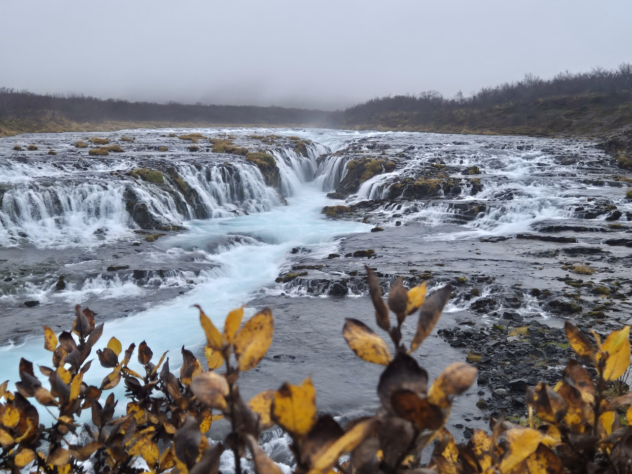 Wide view of Brúarfoss as bright blue glacial water cascades over rocky ledges under a misty sky.