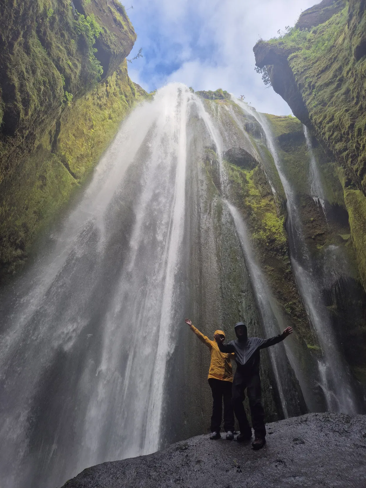 couple pose with arms wide at the bottom of a dramatic Icelandic waterfall, framed by steep green moss-covered canyon walls and bright sky above.