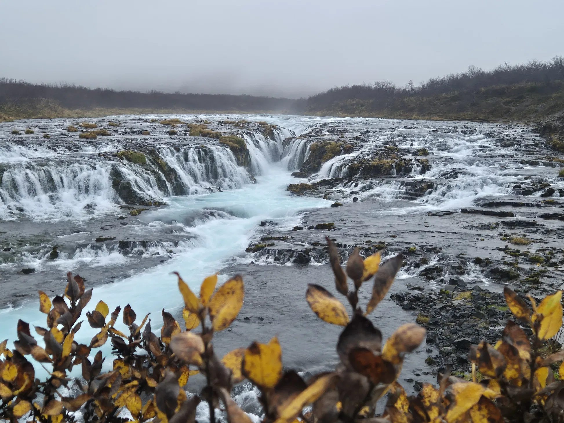 Broad tiered waterfall with milky blue water flowing through dark rocks under a gray sky
