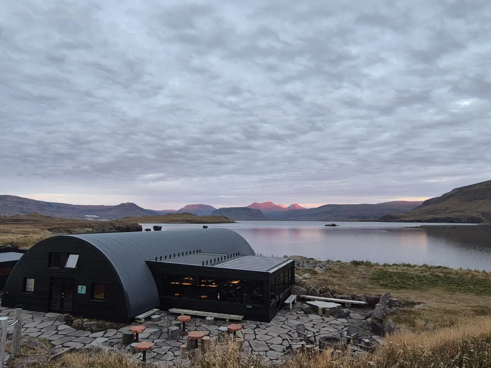 Modern black lodge beside a calm lake with distant mountains under a wide cloudy sky.